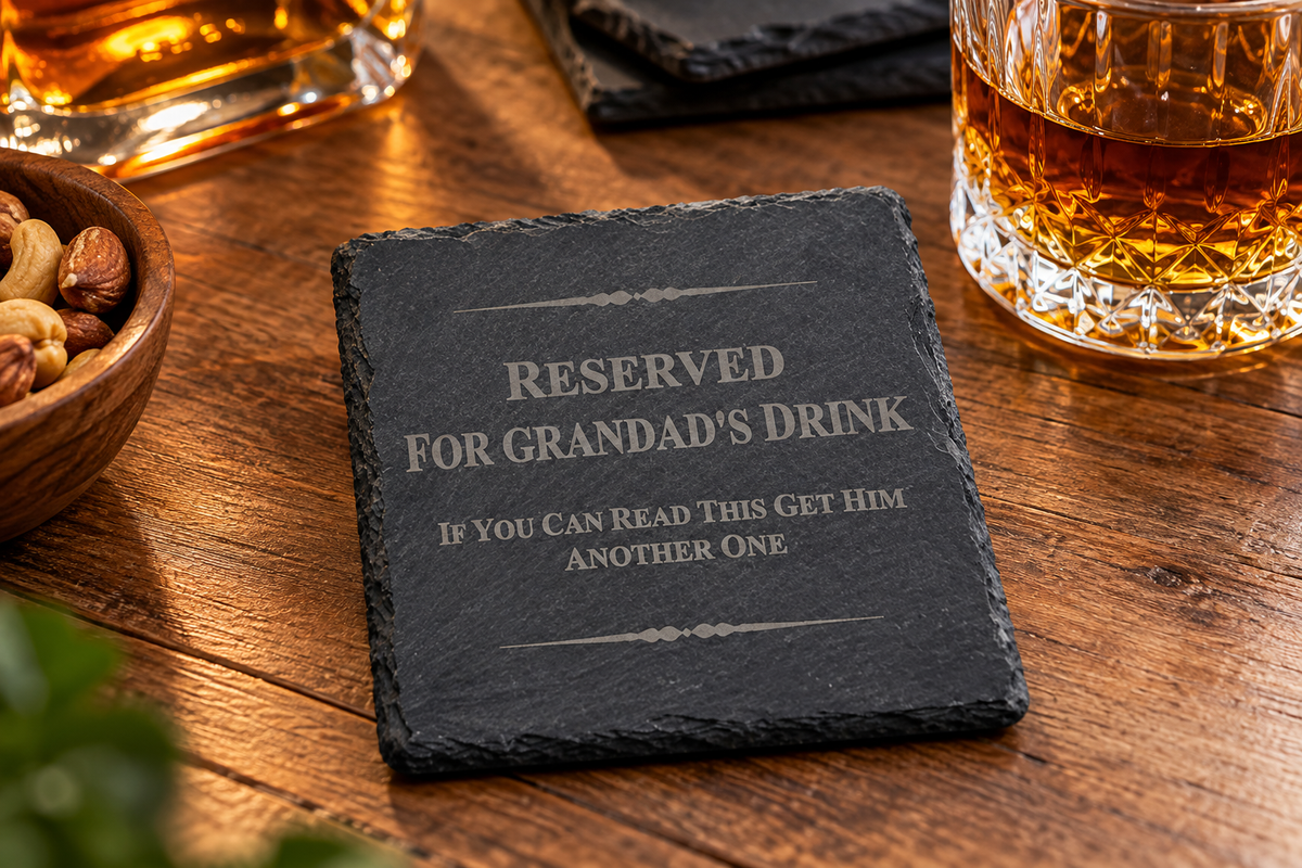 A square dark slate coaster with engraved text reading 'RESERVED FOR GRANDAD'S DRINK IF YOU CAN READ THIS GET HIM ANOTHER ONE' placed on a wooden table with whiskey glasses and nuts nearby.
